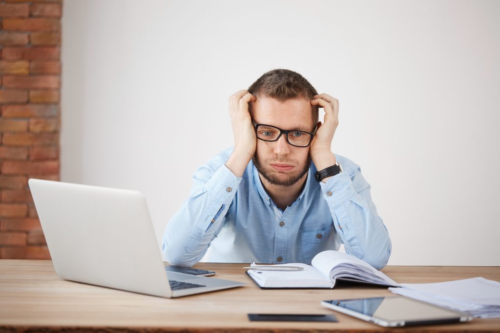 portrait of young unshaven company director in glasses holding head with hands, looking aside with tired and unhappy face expression, being exhausted after hard day at work.